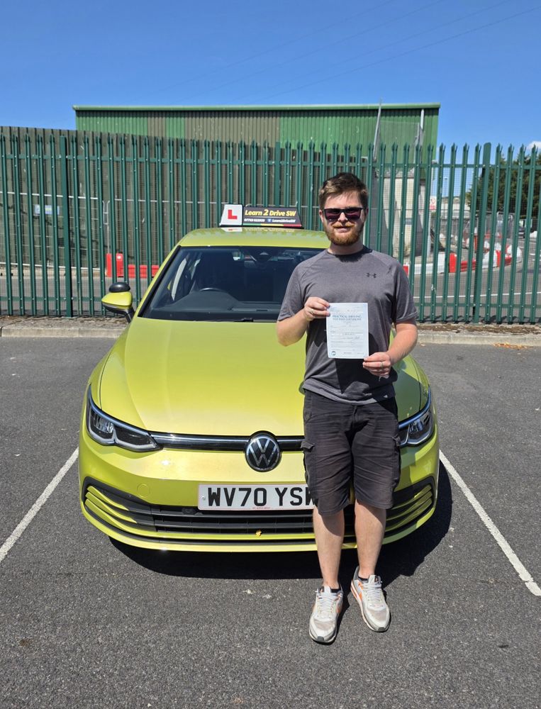 Luke holding his driving test pass certificate in front of the driving school car